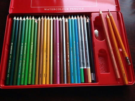50本近い水彩色鉛筆（ファーバーカステル48色セットと他のメーカーの水彩色鉛筆）が赤い金属製の入れ物に収納されている。入れ物の右端には、白い練り消しゴムと橙色の軸のH鉛筆も置かれている