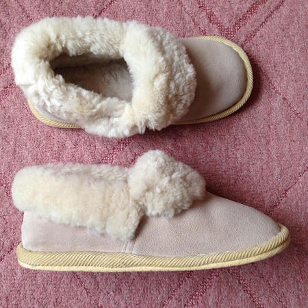 ンク色のカーペットの上に、ベージュ色の冬用スリッパ（室内履き）が置かれている。スリッパの内側は羊の毛（ムートン）で覆われており、もこもことしていて暖かそうに見える。画像上部は左足の靴の俯瞰図、画面下部は右足の靴の側面図