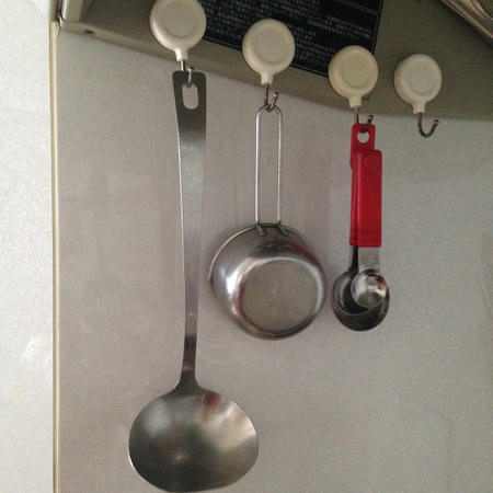 磁石つきの白いマグネットフックが4つ並んでおり、それぞれに金属製のおたまと金属製の計量カップと赤い柄の計量スプーンが掛けられている。背景は台所のコンロ付近の白いつややかな壁